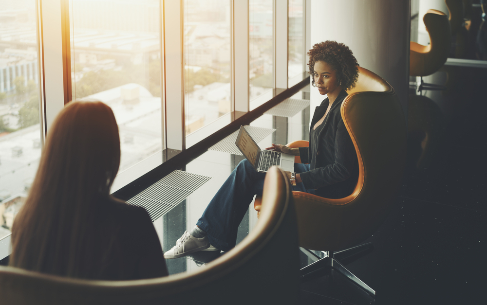 Business meeting two woman: caucasian and afro american with laptop and curly hair, both are sitting in armchairs on high floor of skyscraper near window, office luxury interior with reflections; Shutterstock ID 675616297; PO: 123